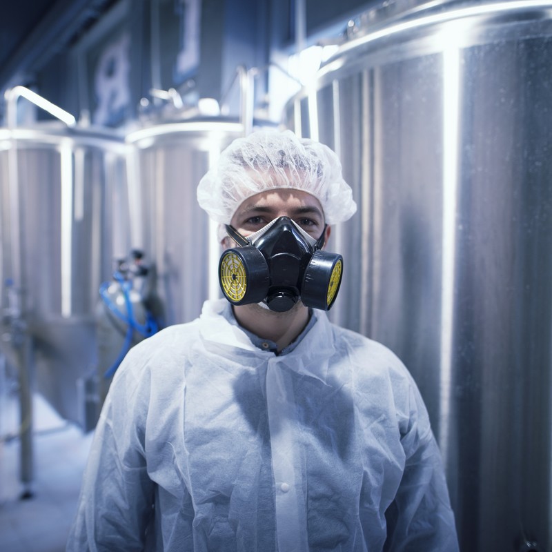 Man in white protective uniform with hairnet and protective mask handling hazardous chemicals.